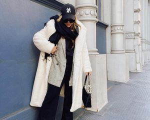 Audree Kate Lopez wears a white fluffy coat, checked blazer, scarf, New York Yankees hat, trousers, and black handbag