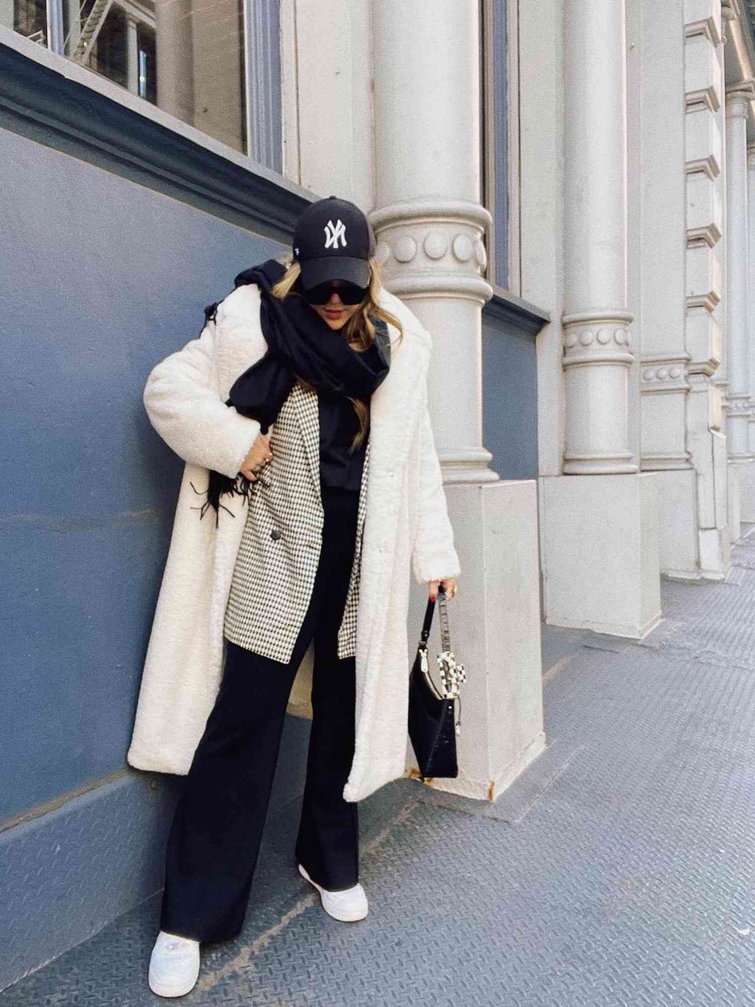 Woman wearing fluffy white coat, checked blazer, black scarf, New York Yankees hat, sunglasses, black trousers, and white sneakers