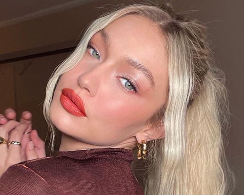 Gigi Hadid wears a radiant, warm-toned makeup look with orange-red lipstick