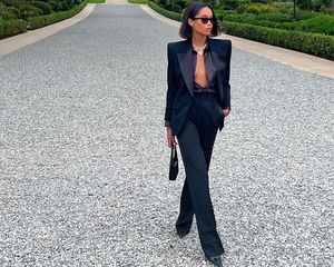 Laura Harrier wears black open button-down, blazer, tailored pants, pointed toe shoes, handbag, and sunglasses