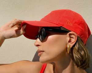 Hailey Bieber wears a red baseball cap, gold earrings, and black sunglasses