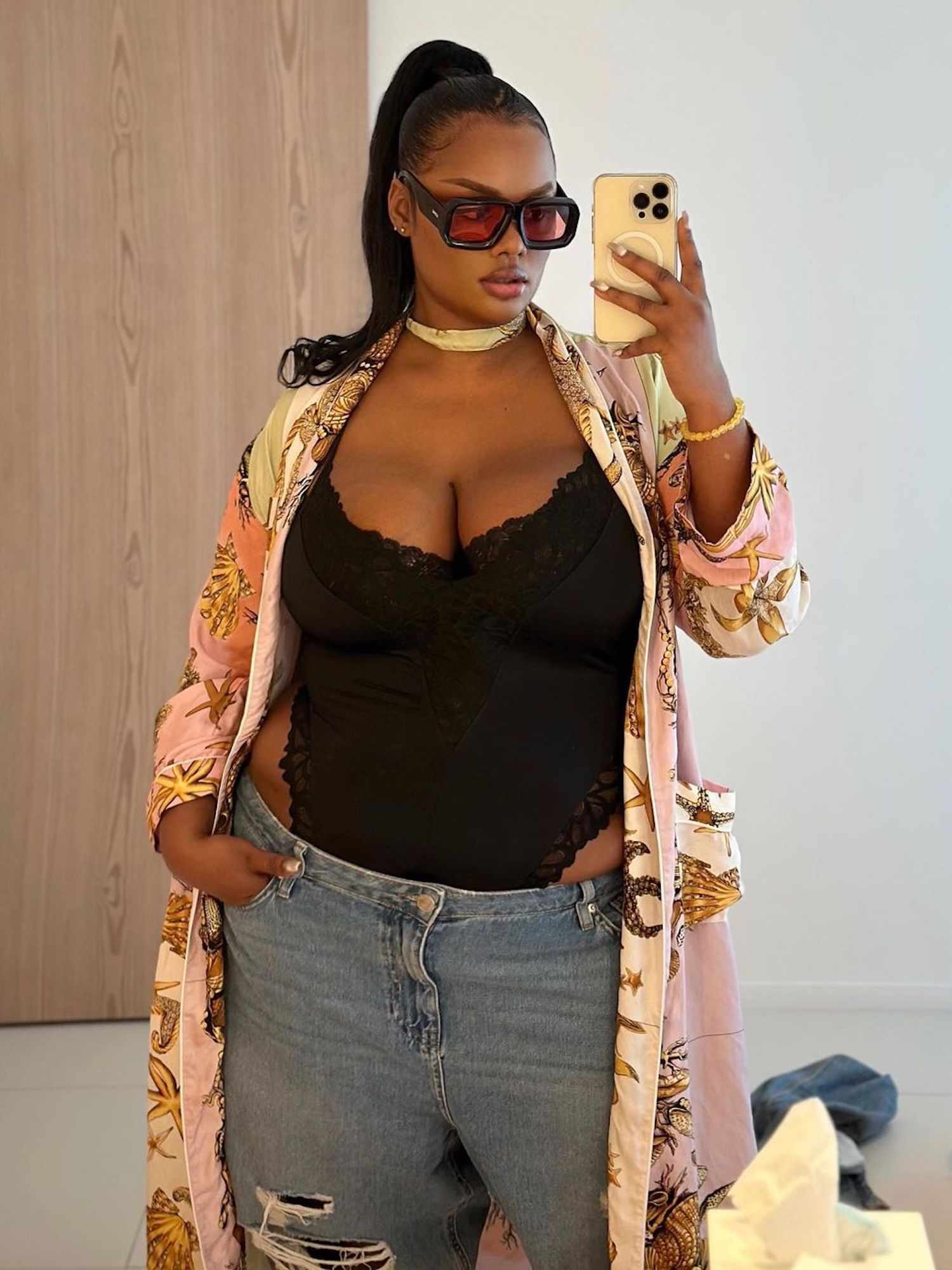 Precious Lee wears a black lace-trim bodysuit, jeans, tinted sunglasses, and printed duster