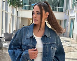 Ashley Graham wears a white ribbed tank top, denim jacket, and high ponytail hairstyle with face-framing pieces