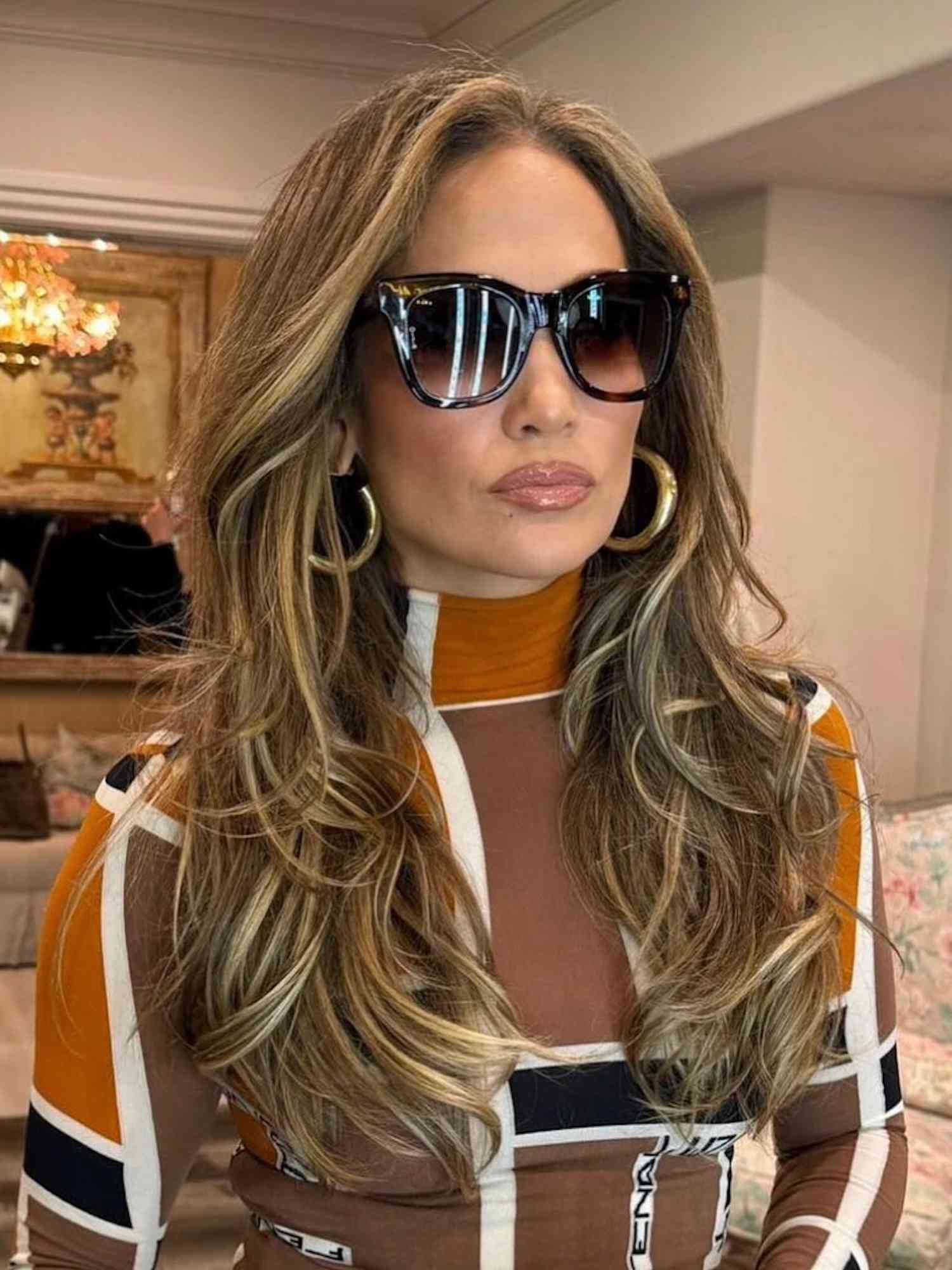 Jennifer Lopez wears oversized sunglasses, gold hoop earrings, and patterned turtleneck top