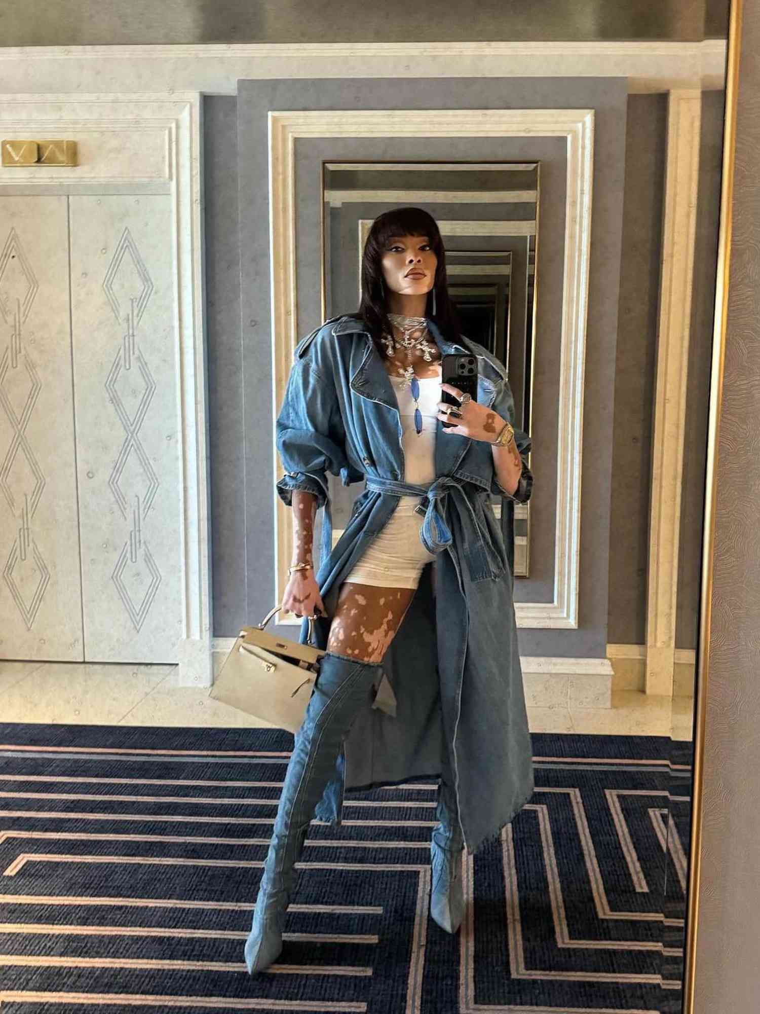 Winnie Harlow wears a light wash denim trench coat, white playsuit romper, and over-the-knee denim boots