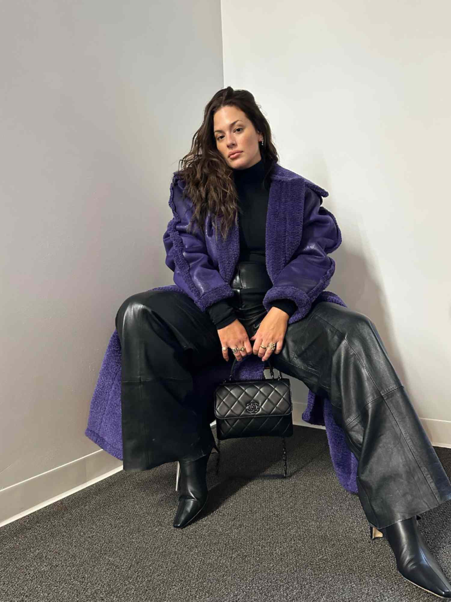 Ashley Graham wearing a purple shearling trim leather coat, black top, leather pants, pointy-toe boots, quilted Chanel bag, and silver jewelry