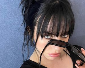 Billie Eilish holding sunglasses with thick black eyeliner, black hair with bangs, and a black t-shirt