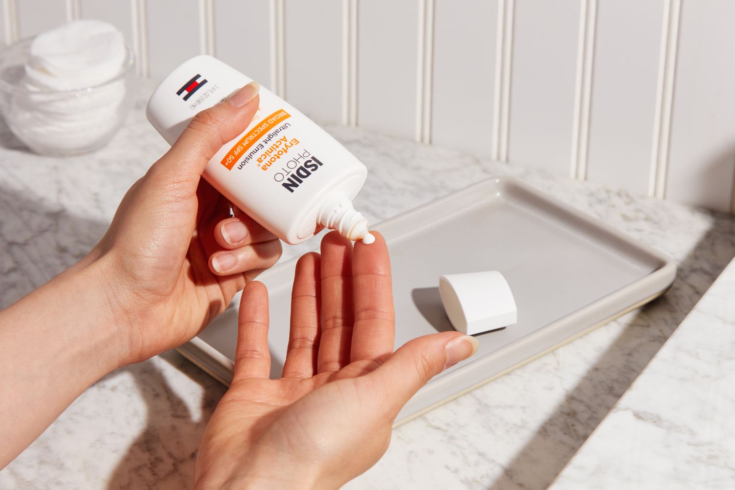 Person dispensing ISDIN Eryfotona Actinica Mineral SPF 50+ Sunscreen on hand over marble counter