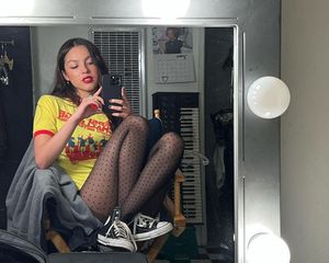 Olivia Rodrigo wtakes a mirror selfie while wearing a colorful graphic tee, sheer dotted tights, and black and white Converse sneakers