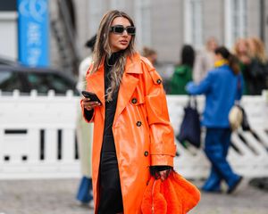 Woman wearing orange trench coat, fuzzy handbag, black outfit, and rectangular sunglasses