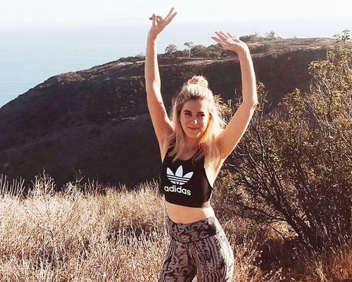 Blonde-haired woman wearing Adidas workout clothing and standing with her hands up