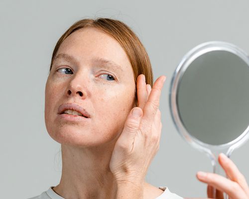 A woman touches her face while looking in a hand mirror