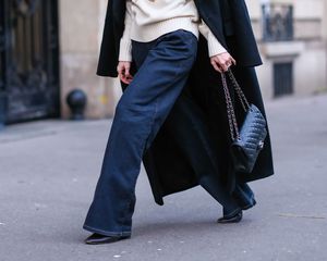 Woman wearing flared wide leg dark wash jeans, white sweater, long black coat, puffy Chanel bag, and black shoes