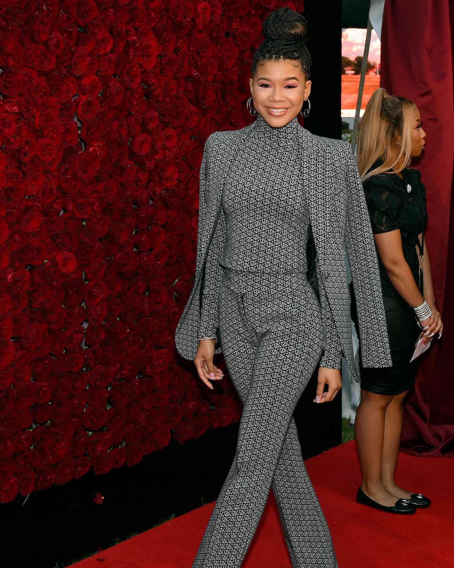 Storm Reid wears a matching black and gray patterned turtleneck, trousers, and suit jacket with oversized hoop earrings and braided topknot