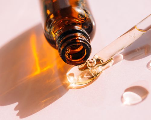 Drops of facial serum pouring out of amber glass bottle nearby pipette on pink background.