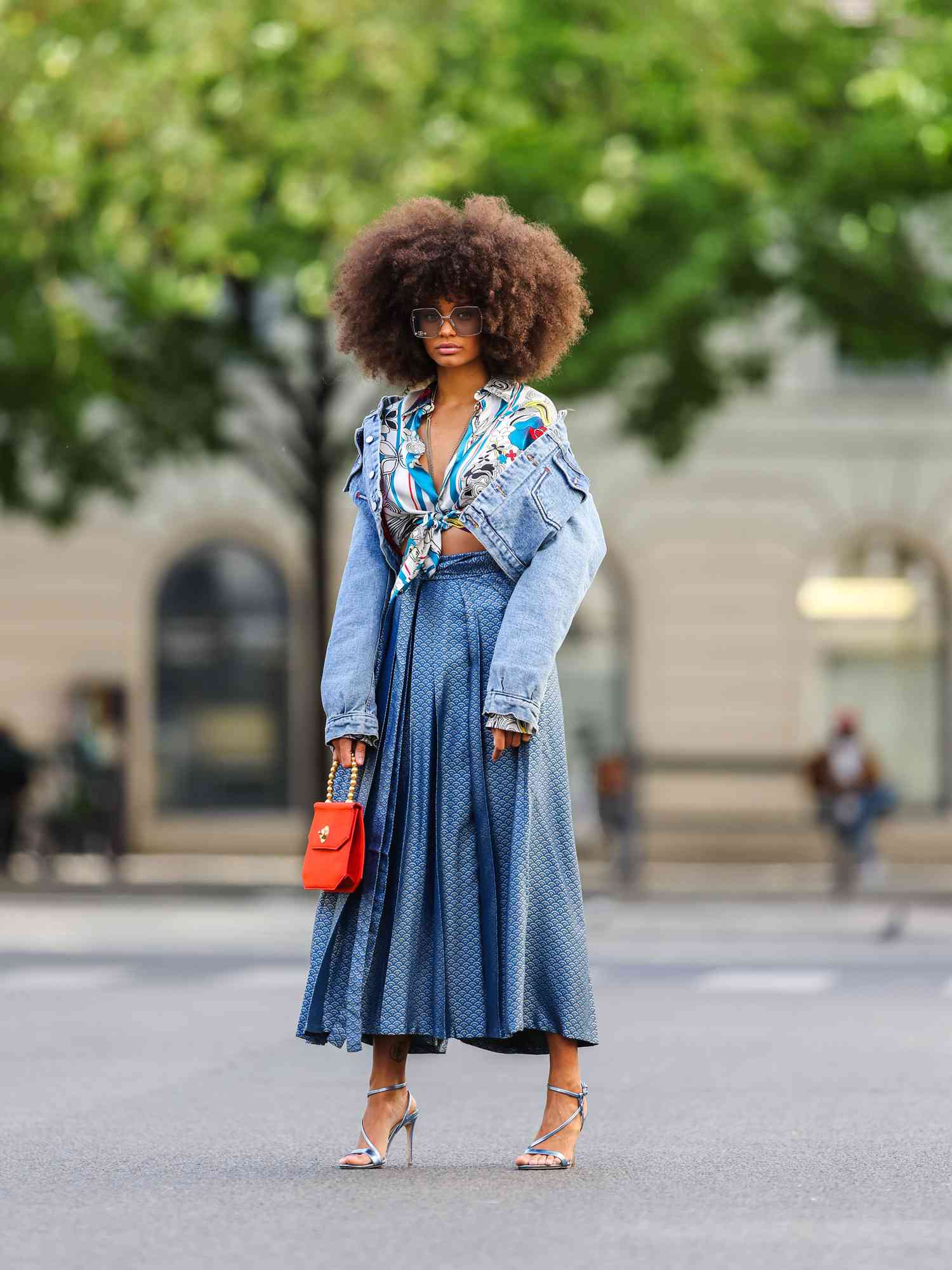 Alicia Aylies wears silver Gucci sunglasses, a silver chain necklace, patterned silk knotted Roberto Cavalli shirt, a blue faded short denim jacket, a blue and silver pattern pleated midi skirt, silver strappy Guess pumps, and a red leather Celine handbag with gold pearl handle