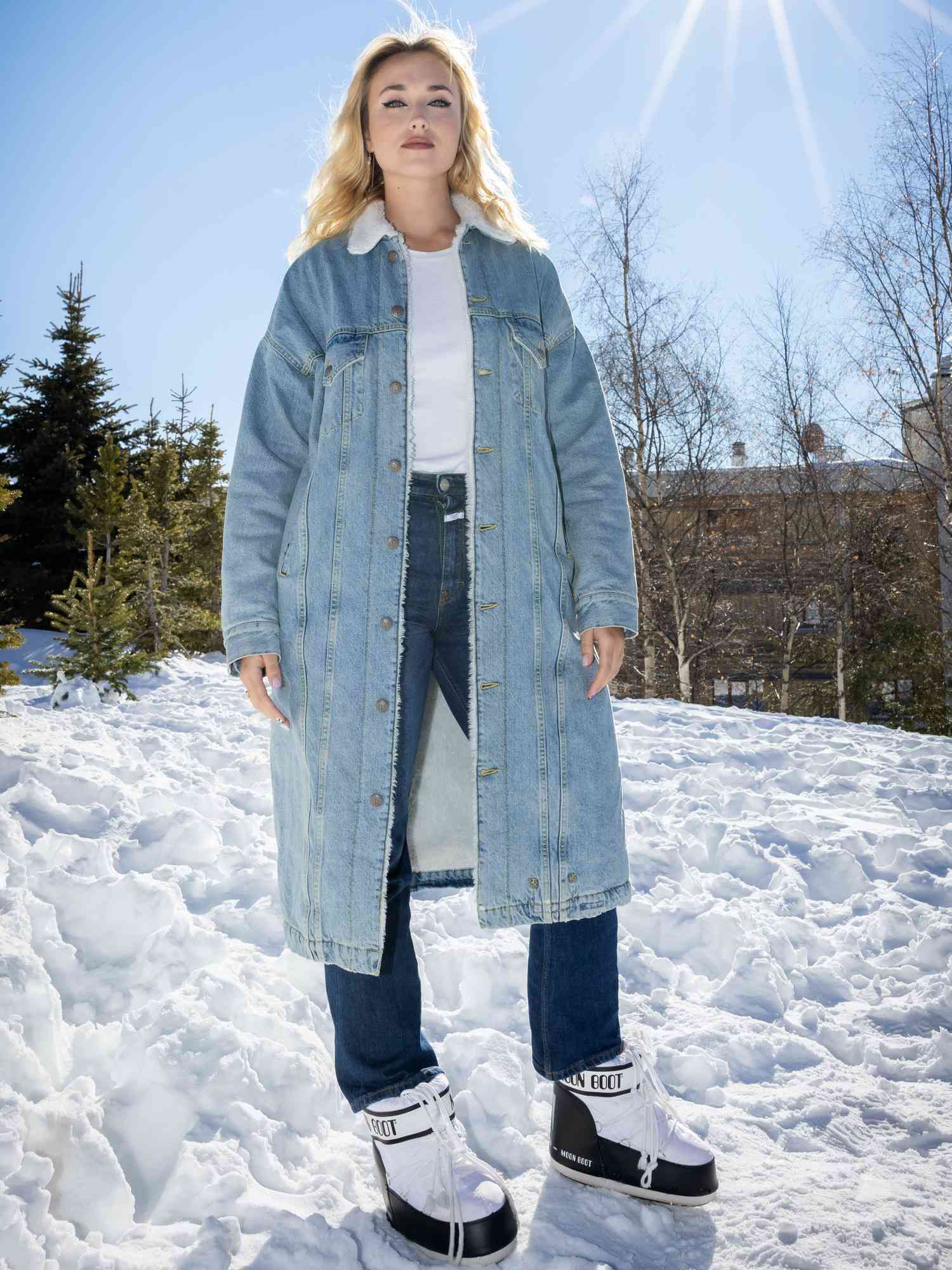 Chloe Jouannet wears a long denim jacket, white tee, dark wash jeans, and white Moon Boots while standing on snowy hill