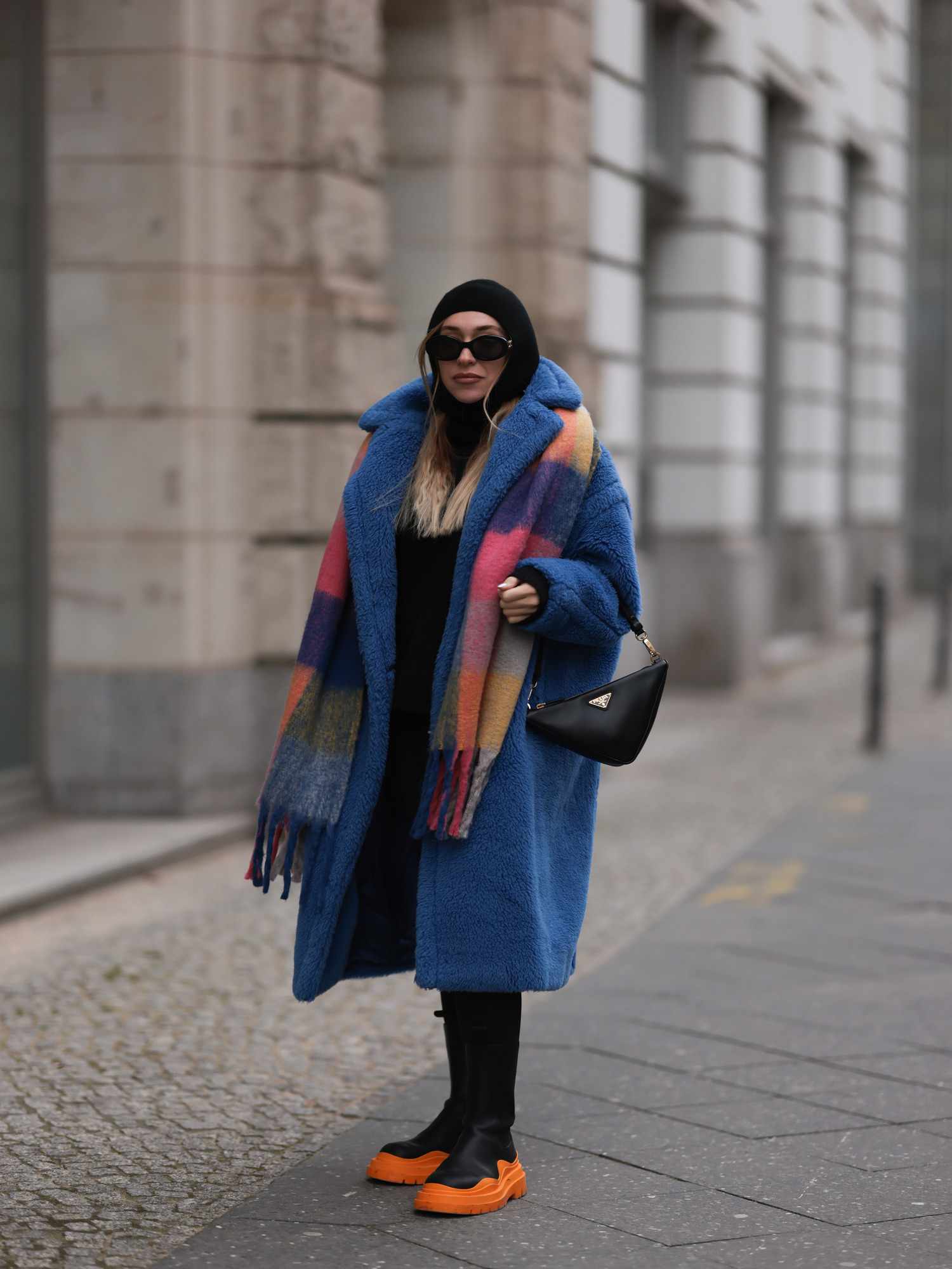 Sonia Lyson wears a triangle black leather Prada bag, black and orange leather boots, blue teddy oversized coat, a colorful checked knit scarf, round black sunglasses, and a black balaclava. 