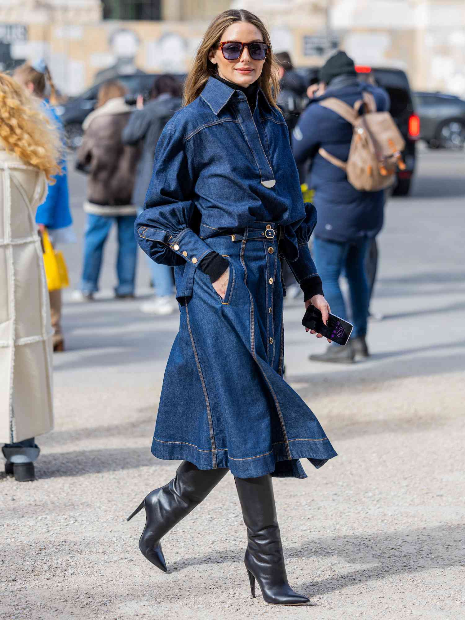 Olivia Palermo wears a denim button-down midi skirt, collared blouse, and black knee-high boots