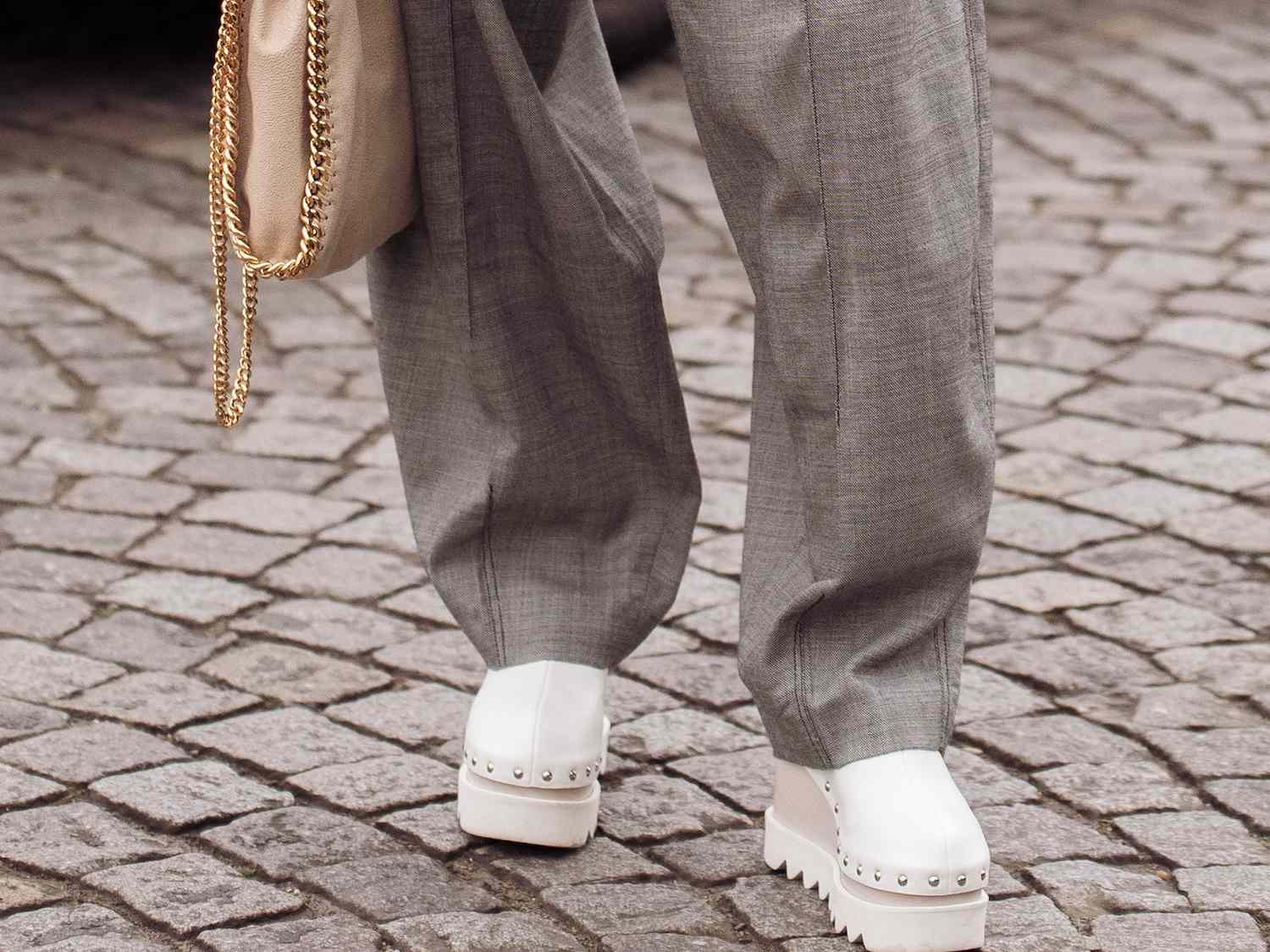 Close-up of woman wearing gray wide leg pants, white platform shoes, and a beige Stella McCartney bag with chain border