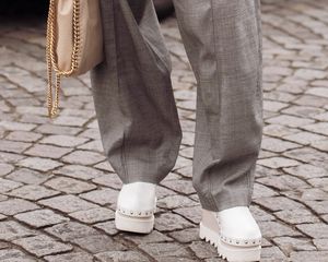 Close-up of woman wearing gray wide leg pants, white platform shoes, and a beige Stella McCartney bag with chain border