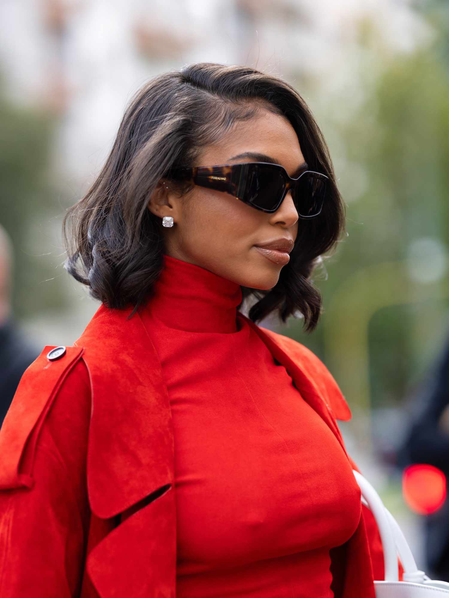 Lori Harvey wears modern tortoiseshell sunglasses, a bob hairstyle, and a red turtleneck and trench coat