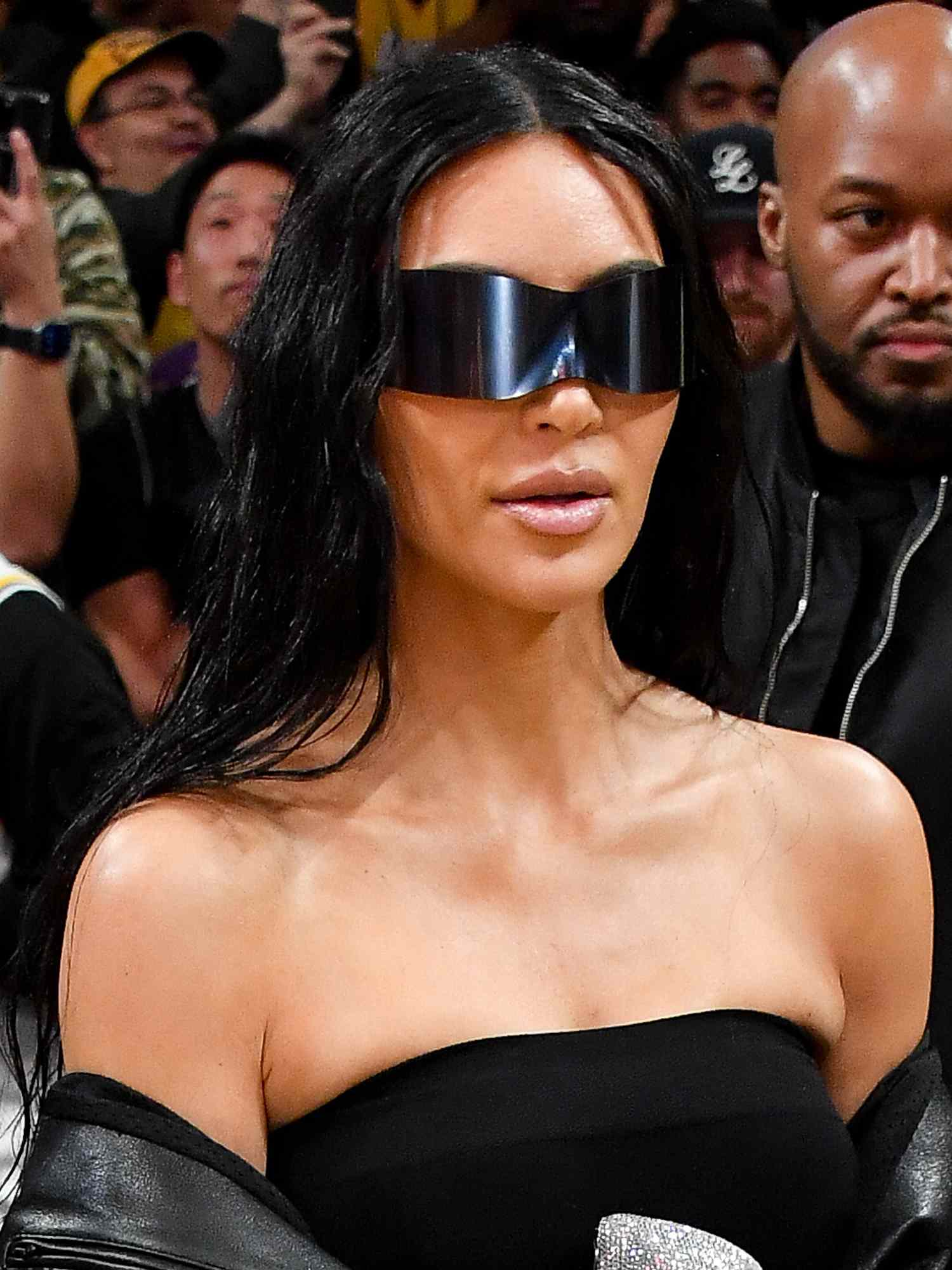 Kim Kardashian wears black shield sunglasses, a strapless black top, and a leather jacket