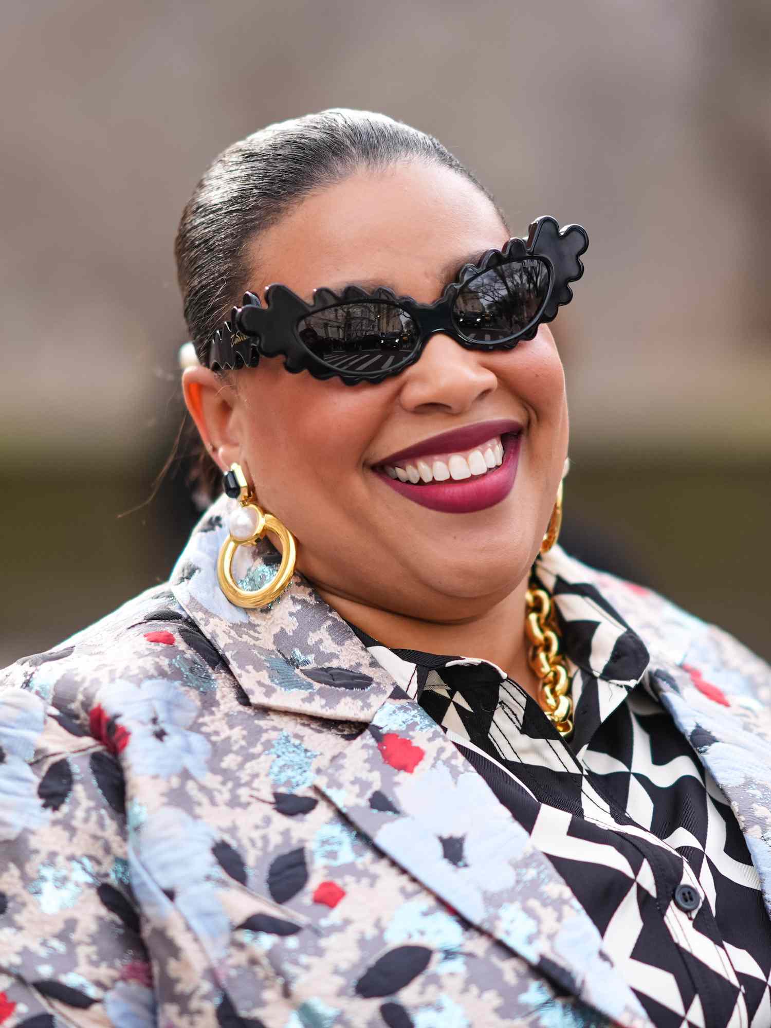 Greivy wears black cat-eye sunglasses with scalloped frames, gold jewelry, and colorful patterned dress and coat