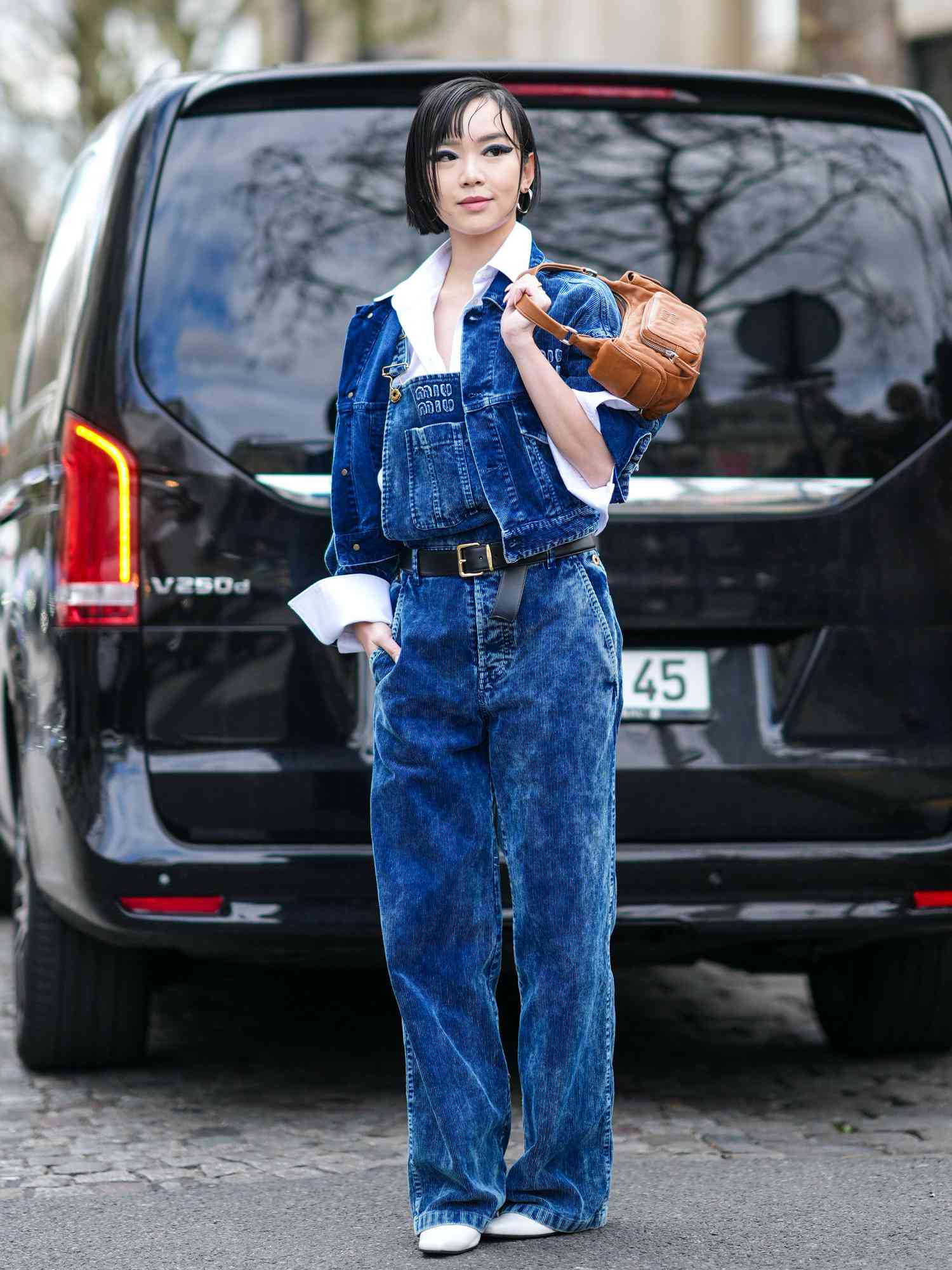 Woman wearing a blue denim jacket, denim overalls, white pointed shoes, and a brown leather bag