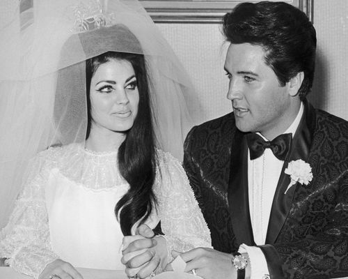 Priscilla Presley wears a puff-sleeved white gown, wedding veil, and crown while sitting with husband Elvis