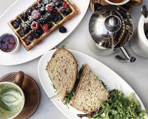 Table top with waffles and berries; sandwich halves with salad; matcha cup; tea cup; and tea kettle