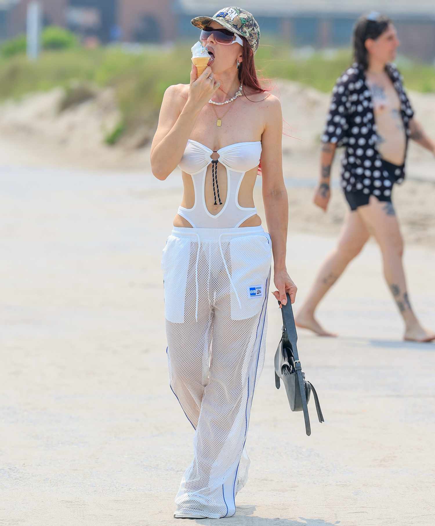 Julia fox at the beach eating an ice cream cone wearing an army hat, sunglasses, white monokini, and white mesh trousers