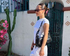 Woman carrying black Marge Sherwood handbag and wearing white outfit, black oval sunglasses, and slicked back bun