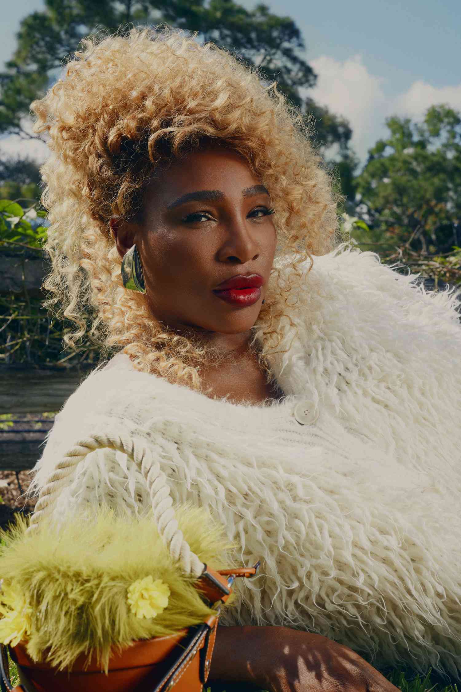 Serena Williams laying down on grass in a white fluffy sweater with curly blonde hair