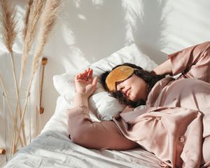 Woman sleeping with eye mask and button-down pajamas in bed next to vase of pampas grass