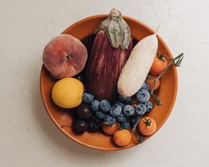 An orange bowl of produce, including blueberries, nectarines, an egglplant, and cherries, on a cream colored surface.