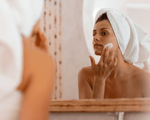 A woman looking in the mirror with her hair in a towel and applying a face mask