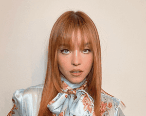 Sydney Sweeney with straight copper hair, standing against a beige wall