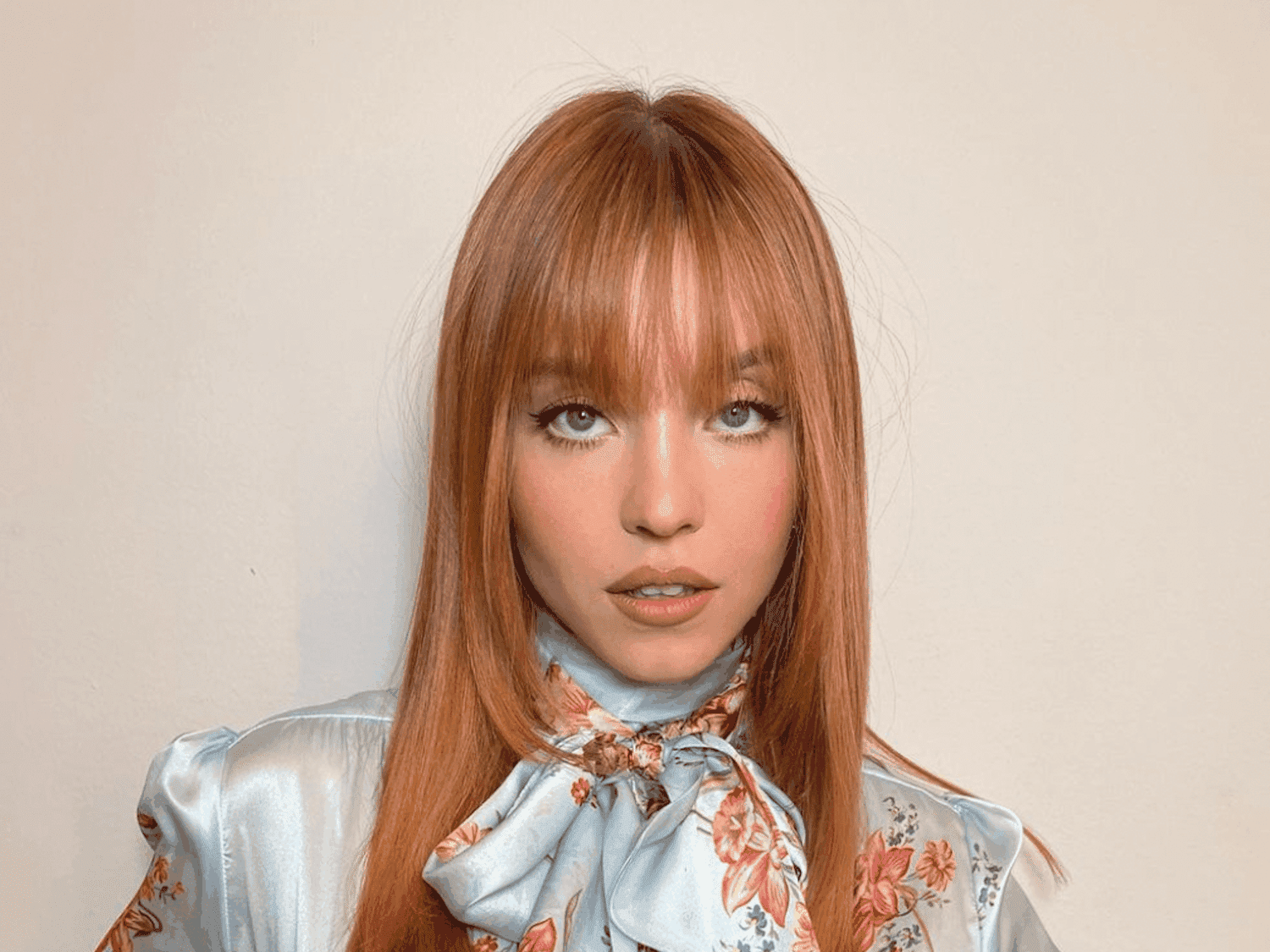 Sydney Sweeney with straight copper hair, standing against a beige wall