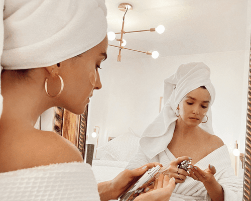 A woman in a robe and her hair in a towel looking in the mirror and applying skincare