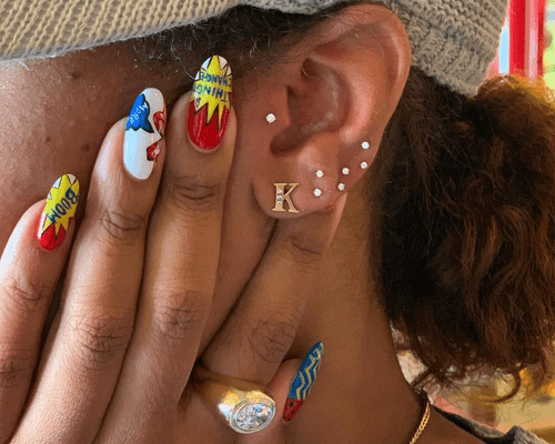 A woman's ear with multiple piercings and her hand on her face with cartoon design nails