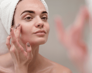 A woman with her hair in a towel, looking in the mirror and touching her face