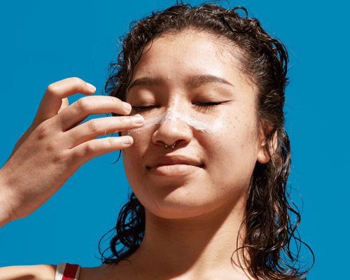 Woman in front of a blue background applying sunscreen to nose and cheeks.