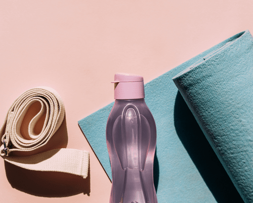 A pink water bottle next to a blue yoga mat and nude yoga strap on a pink background.